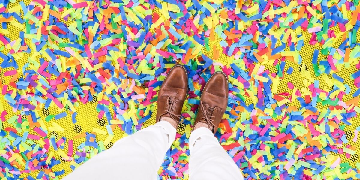 Brown dress shoes standing on a yellow surface with colorful confetti spread everywhere